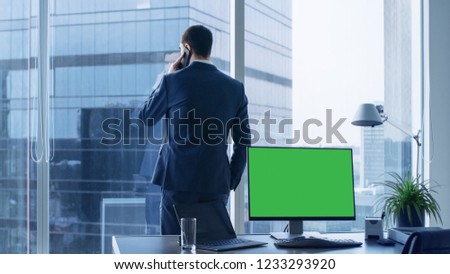 Confident Businessman in His Office Making Phone Call, Personal Computer on His Desk Shows Green Chroma Key Screen. In the Window Panoramic Big City View.