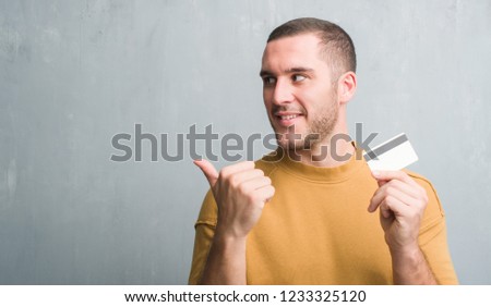 Young caucasian man over grey grunge wall holding credit card pointing and showing with thumb up to the side with happy face smiling