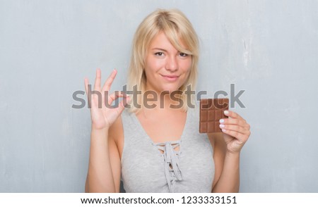 Caucasian adult woman over grey grunge wall eating chocolate bar doing ok sign with fingers, excellent symbol