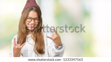 Young beautiful brunette hipster woman wearing glasses and winter hat over isolated background shouting with crazy expression doing rock symbol with hands up. Music star. Heavy concept.