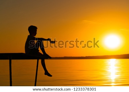 In the evening a small boy sits on the bridge over the calm river with a paper boat. A child is watching a beautiful sunset in summer . Origami.