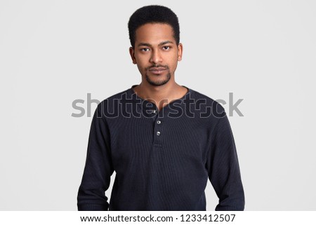 Serious Afro American man dressed in casual sweater, has self confident expression, looks directly at camera, isolated over white background. Ethnic male poses at studio. Facial expressions concept Royalty-Free Stock Photo #1233412507
