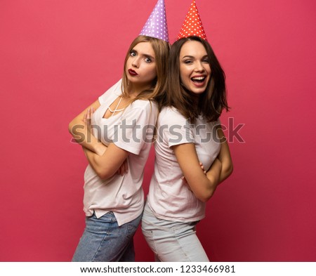 Close up portrait of blonde and brunette young women with birthday hats having fun isolated on pink background.