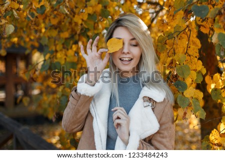 Pretty young happy girl model with blue eyes in a fashion jacket with fur covered her face with a yellow autumn leaf in the gold fall park . Beautiful smiling woman.