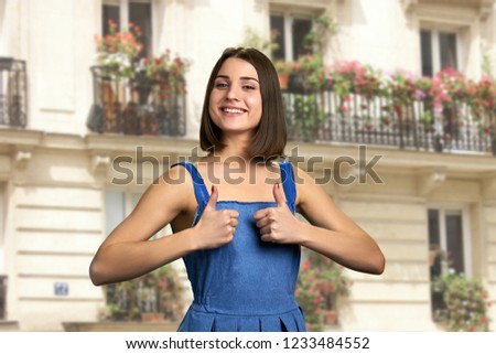 Stylish girl gesturing thumbs up outdoor. Happy beautiful girl in showing thumb up symbol by two hands on buildings background. Summer holidays and tourism concept.