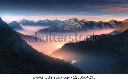 Mountains in fog at beautiful night in autumn in Dolomites, Italy. Landscape with alpine mountain valley, low clouds, forest, colorful sky with stars, city illumination at dusk. Aerial. Passo Giau Royalty-Free Stock Photo #1233564235