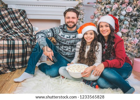 Picture of family sitting on floor and smiling on camera. Young man has remote control in hand. Girl holds bowl of popcorn. She leans to mother.