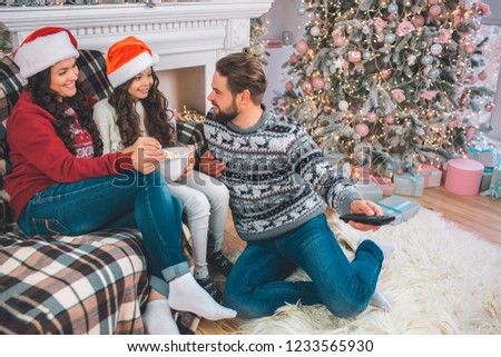 Lovely picture of family sitting together on gfloor. Girl holds bowl of popcorn. Father looks at her and uses remote control at the same time. Young woman and girl wears Christmas hats.