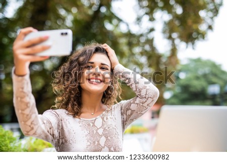 Portrait of happy young girl taking selfie with mobile phone while sitting with laptop computer at a cafe outdoors