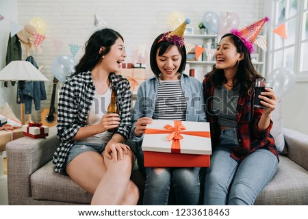 three friends sharing happy moments opening birthday present. young asian girl curiously looking inside gift box. beautiful ladies giving surprise and drinking beer love alcohol.