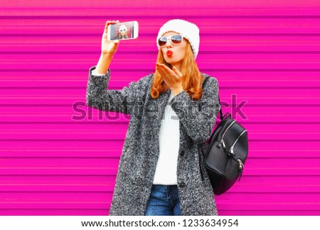 Pretty young woman taking selfie by smartphone blowing red lips sends an air kiss in coat jacket, white knitted hat on colorful pink wall background