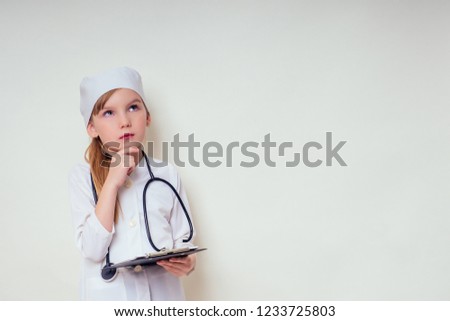 smiling little girl in doctor uniform with medical tools stethoscope writing something to clipboard on white background in studio copy space.future profession vocational guidance career