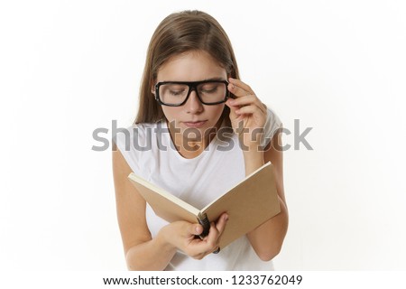 Hardworking straight A-student girl wearing t-shirt and eyeglasses for good vision, holding textbook in hands, reading something, having concentrated facial expression. Learning and knowledge