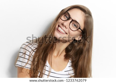Playful teenage girl in eyewear fooling around, looking at camera and sticking out her tongue as if teasing you. Childish female student posing in studio, showing tongue at camera, having good mood