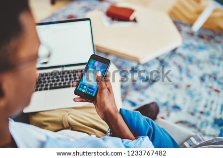 Cropped image of dark skinned male using smartphone for networking via wifi connection at home checking mail, hipster guy holding mobile phone share multimedia content synchronize with laptop computer