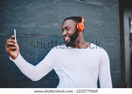 Cheerful african american guy in headphones spending time outdoors with modern gadgets taking picture on smartphone camera, smiling dark-skinned male hipster making selfie while listening music
