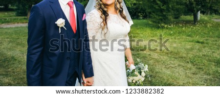 Wedding couple walking in the green park. Curvy bride in white lace dress and groom are holding hands. Overweight happy people. Love story outdoors. Beautiful bouquet.