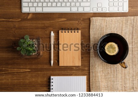 A cup of tea with lemon, a notebook, a pen, a flower, a book, a white keyboard on a natural wooden background