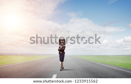 Business woman in suit with an old TV instead of head keeping arms crossed while standing on the road with beautiful landscape on background.