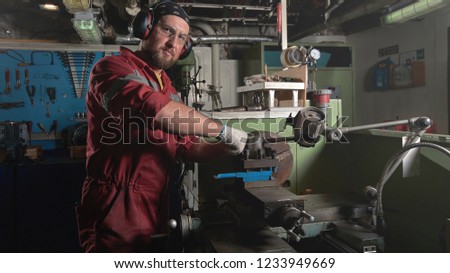 Worker in red uniform operating in manual lathe in metal big workshop with dark environment.