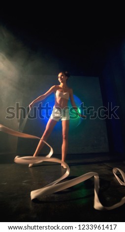 Long shot of an young gymnast woman with a slim sport body training with a gymnastics tape against the black wall with a big fan lit with blue and green neon light.