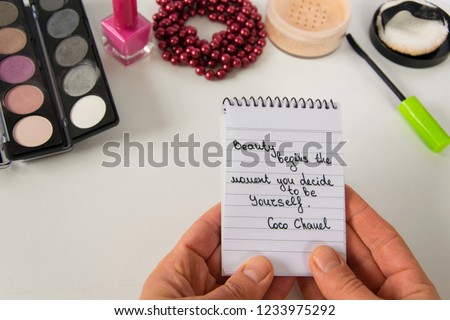 Woman hand holds Coco Chanel quotes written on a block note, pearl accessories and make up on white background, inspiration phrase "Beauty begins the moment you decide to be Yourself"
