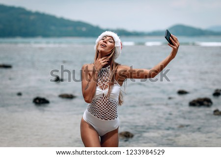Summer beach vacation girl in santa hat taking fun mobile selfie photo with smartphone. Girl wearing white swimsuit posing for selfie.