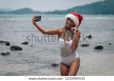 Summer beach vacation girl in santa hat taking fun mobile selfie photo with smartphone. Girl wearing white swimsuit posing for selfie.