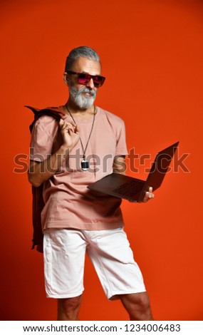 Portrait of cool, manly, fashionable old man senior male with thick gray beard using laptop computer, standing at full height and looking at the picture in the studio