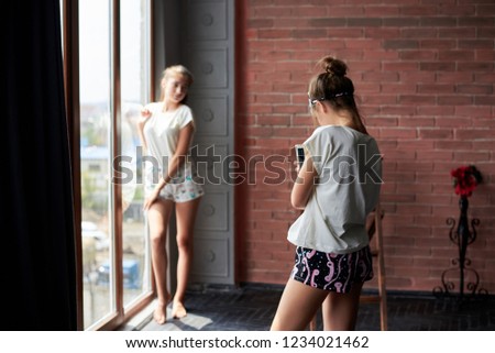 Young girl is taking picture with her phone of her friend standing by the window near brick wall wearing pajamas