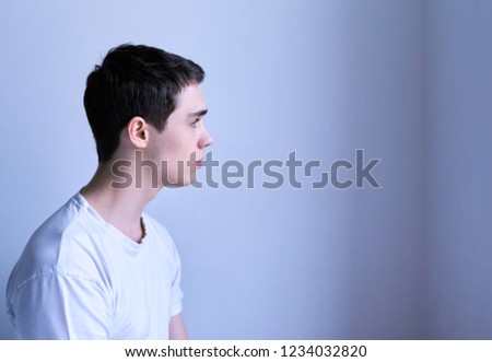 A young man sits in the white shirt on the white background of the wall. Guy looks out the window. Side view.