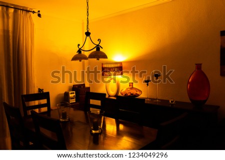 Interior of dining room with vintage lamp, glass vase and candlesticks on chest of drawers, wooden table and chairs in night-time lighting