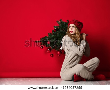 Young woman hold winter christmas fir tree decoration in knitted hat under snow. New year concept on red background