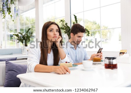 Bored young girl sitting at the cafe table while her boyfriend using mobile phone Royalty-Free Stock Photo #1234062967