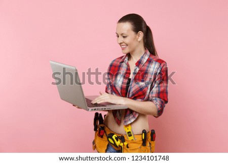 Young happy handyman woman user in plaid shirt, denim shorts, kit tools belt full of instruments working on laptop pc computer isolated on pink background. Female doing male work. Renovation concept