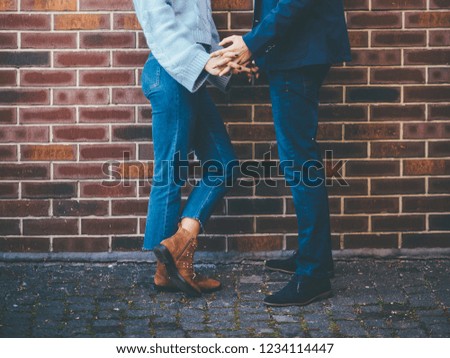 Cropped photo of a stylish couple - they hold hands near dark red brick wall