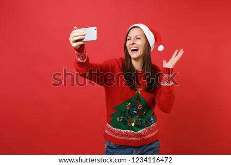 Laughing Santa girl in Christmas hat doing taking selfie shot on mobile phone, spreading hands isolated on red background. Happy New Year 2019 celebration holiday party concept. Mock up copy space