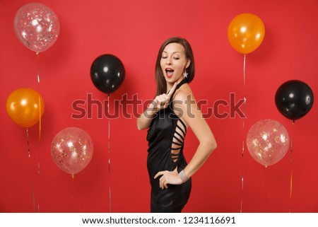 Attractive woman in black dress celebrating, looking behind herself, pointing finger on jewelry on bright red background air balloons. Women's Day Happy New Year birthday mockup holiday party concept