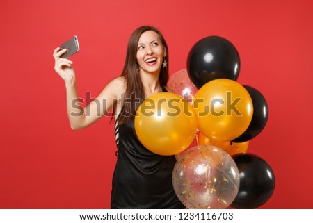 Dreamy woman in black dress celebrating holding air balloons doing taking selfie shot on mobile phone isolated on red background. Valentine's Day Happy New Year birthday mockup holiday party concept