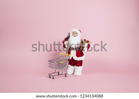 a santa claus figurine plush placed in front of a shopping trolleyon a pink background. Color harmony. Minimal still life color photography