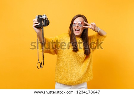 Excited young woman in heart eyeglasses doing taking selfie shot on retro vintage photo camera showing victory sign isolated on yellow background. People sincere emotions, lifestyle. Advertising area