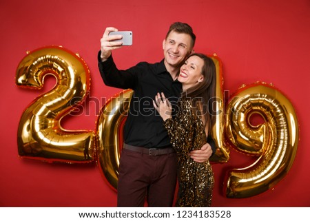 Couple guy girl in dress black shirt celebrating holiday party hold cellphone isolated on bright red wall background golden numbers air balloons studio portrait. Happy New Year 2019 Christmas concept
