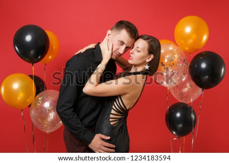 Stunning young couple in black clothes celebrating birthday holiday party isolated on bright red background air balloons. St. Valentine's International Women's Day Happy New Year 2019 concept.