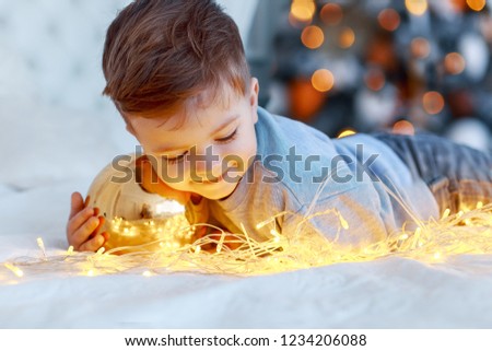 A a cute little boy with a christmass toy ball in front of the christmas tree on the bed with candles and gifts. New year's eve. Cozy holiday at the fir-tree. love, happiness and family concept