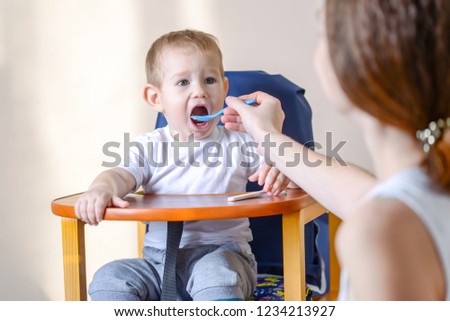 Little baby boy eats opening his mouth wide sitting on a chair in the kitchen. Mom feeds holding in hand a spoon of porridge