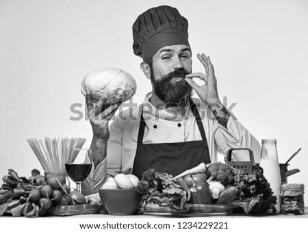 Chef prepares meal showing Perfect sign. Cook with happy face in uniform sits by table with vegetables and kitchenware. Restaurant cuisine concept. Man with beard holds cabbage on white background