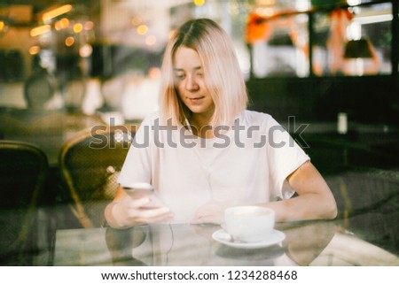 A young girl wearing white t-shirt is sitting in a cafe with a big cup of coffee or tea and looking into her smartphone behind a glass vitrine