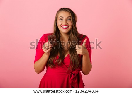 Image of a pretty hungry young woman isolated over pink wall background holding chocolate showing thumbs up.