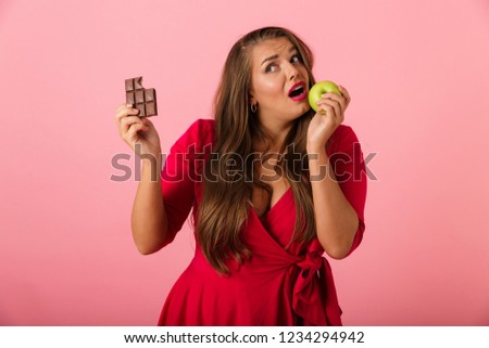 Image of a beautiful hungry young woman isolated over pink wall background holding chocolate and apple.