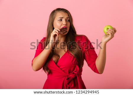 Image of a confused young woman isolated over pink wall background holding chocolate and apple.